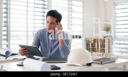 Thoughtful Asian man using digital tablet considering architectural design details at a contemporary workspace with building model and blueprints Stock Photo