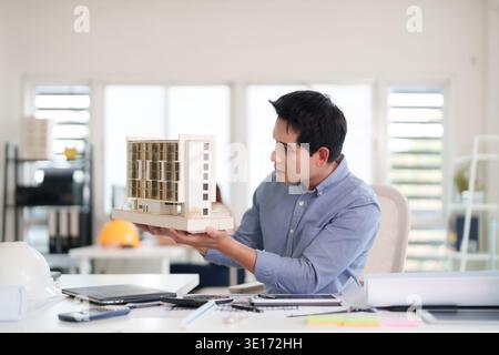 Dedicated Asian adult male architect analyzing intricate scale model for innovative construction. Focused on future urban planning. Stock Photo