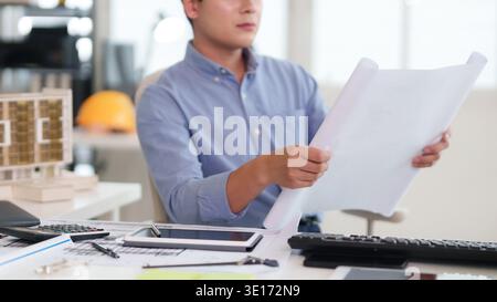 Focused Asian male professional examines architectural drawings. Desk features building model, calculator, tablet. Modern office environment design co Stock Photo