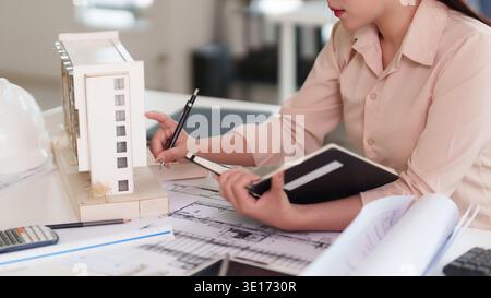 Adult Asian woman designs architectural blueprint plans builds building model uses drafting tools for innovative urban property development Stock Photo