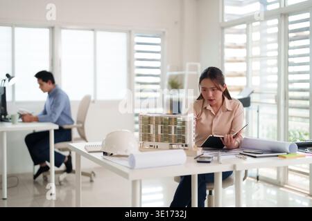 Asian adult female designer diligently inspects intricate building model. Her male coworker works nearby in a contemporary bright office setting. Stock Photo