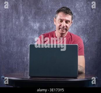 Man sitting in front of a laptop. The man is of Caucasian origin, forty years old, and wears a burgundy T-shirt Stock Photo