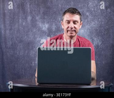 Man sitting in front of a laptop. The man is of Caucasian origin, forty years old, and wears a burgundy T-shirt Stock Photo