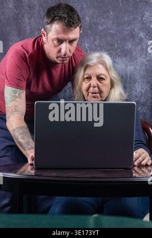 A forty-year-old man teaches his seventy-year-old mother how to use a computer. The man wears a burgundy T-shirt while the woman has long blonde hair Stock Photo