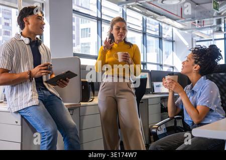 Diverse coworkers chatting, holding coffee mugs and tablet near large windows in modern office. Collaboration, teamwork, headphones, monitors, openplan, cityscape, daylight || Model released Stock Photo
