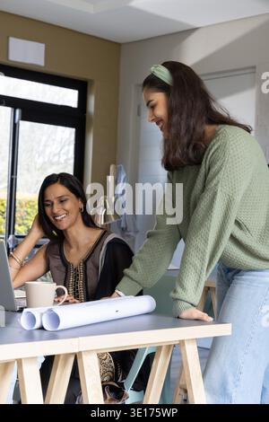 Diverse female colleagues collaborating on laptop with plans in bright home studio, wearing tunic. Teamwork, blueprints, computer, outdoors, nature, design, creativity || Model released Stock Photo
