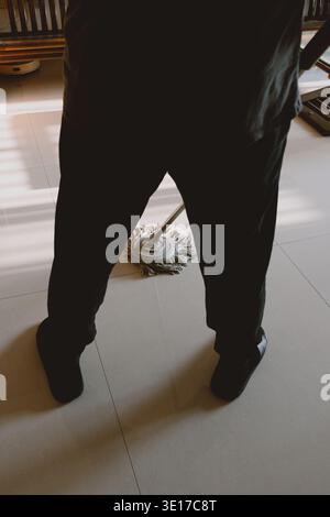Person in dark pants mopping tiled floor indoors with sunlight streaming through window in modern Stock Photo