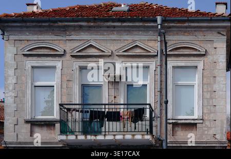 Historic stone building with two windows and a balcony displaying laundry, featuring an air conditioning unit and a red-tiled roof under a clear blue Stock Photo