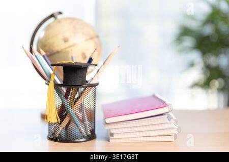 Stack of textbooks with graduation cap and red tassel beside pencil holder and globe on desk in bright modern room. Education, study, elearning, acade Stock Photo