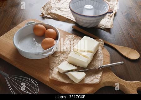 Baking ingredients with sliced butter and fresh eggs on wooden board. High angle shot of farmhouse kitchen table featuring sliced butter on parchment Stock Photo
