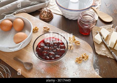 Baking ingredients with canned cherries, walnuts and fresh eggs on floured wooden board. High angle shot of rustic kitchen table featuring bowl of red Stock Photo