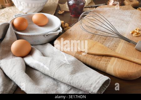 Baking preparation with brown eggs, whisk and wooden spoon on floured board. Close-up shot of rustic kitchen scene featuring fresh eggs in ceramic Stock Photo