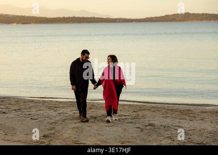 A couple strolls hand in hand on a serene beach, enjoying the tranquil atmosphere by the sea. The calm waters and gentle breeze create a perfect setti Stock Photo