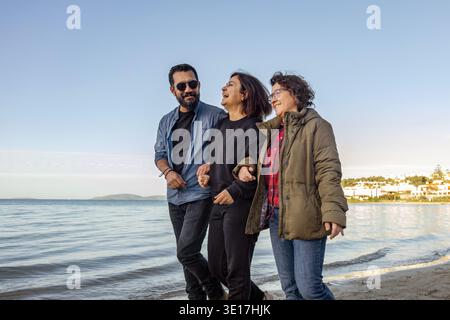 Three friends walk arm in arm along a serene seaside, laughing and enjoying the sunny day. The peaceful atmosphere and gentle waves create a perfect m Stock Photo