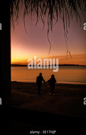 A romantic scene capturing a couple's silhouette as they walk hand in hand by the sea during a beautiful sunset. The sky is aglow with warm hues, crea Stock Photo
