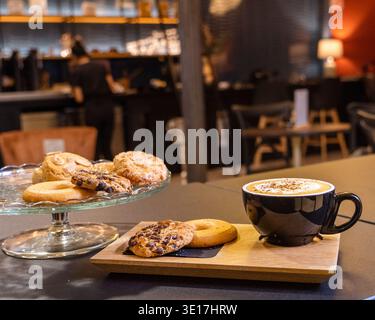 A warm cafe setting featuring a freshly brewed coffee in a black mug on a wooden tray, accompanied by an assortment of cookies on a glass platter, cre Stock Photo