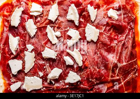 A close-up view of thinly sliced cured meat topped with small chunks of creamy cheese, arranged on a rustic surface, highlighting texture and detail p Stock Photo