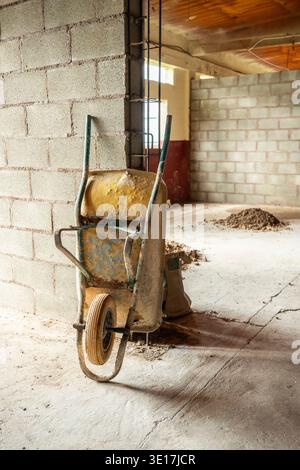 An empty construction site features a worn wheelbarrow leaning against an unfinished brick wall. Piles of dirt highlight ongoing construction activity Stock Photo