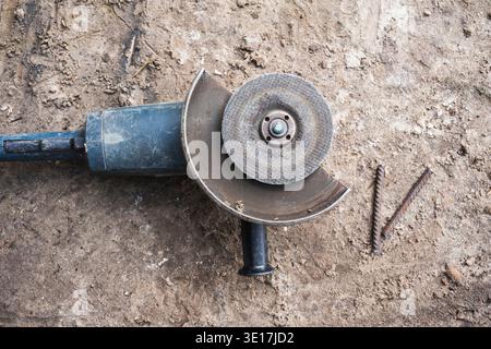 A close-up image of an electric angle grinder resting on a dusty construction site ground. The tool is next to metal rods, illustrating industrial wor Stock Photo