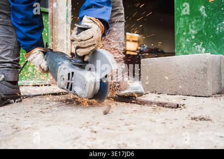 A construction worker in protective gear uses an angle grinder to cut metal rods outdoors. Sparks fly in all directions, emphasizing the intensity and Stock Photo