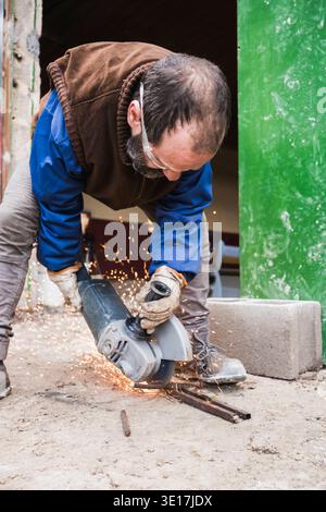 A worker wearing protective gear uses an angle grinder to cut metal rods, creating bright sparks in an outdoor setting. The focus is on safety and man Stock Photo