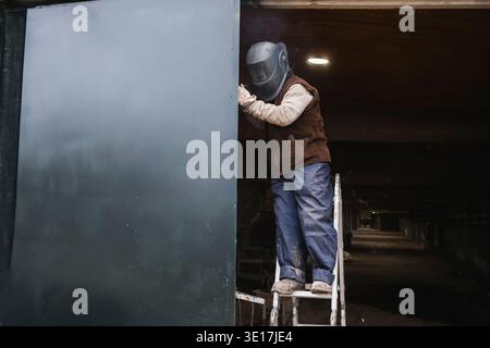 A worker in protective gear welds a large metal structure while standing on a ladder in an industrial setting. The scene highlights industrial labor a Stock Photo