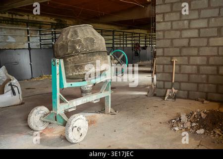 Inside a partially constructed building, a cement mixer and various tools, including shovels, lie on a dusty floor, showcasing ongoing construction an Stock Photo