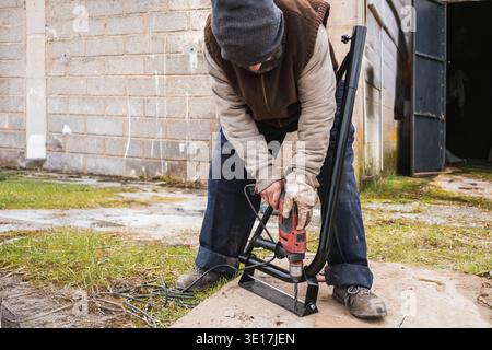 A construction worker is seen using a power drill outdoors, focusing on metalwork. The scene showcases industrial equipment and protective gear in a r Stock Photo