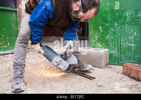 A worker is using an angle grinder to cut metal rods outdoors. The protective gear ensures safety as sparks fly around. The setting is a construction Stock Photo