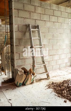 Image of a construction site featuring a concrete block wall with a metal ladder leaning against it. Bags of cement and rubble are visible on the grou Stock Photo