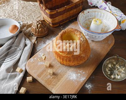 Freshly baked homemade bundt cake on wooden board in rustic kitchen. High angle shot of golden sponge cake on floured cutting board, surrounded by Stock Photo
