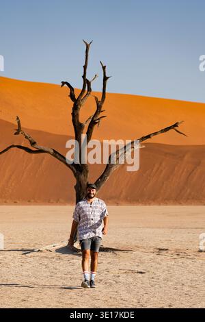A solitary traveler walks through the stark landscape of Deadvlei, Namibia, with a lone dead tree and vibrant orange sand dunes under a clear blue sky Stock Photo