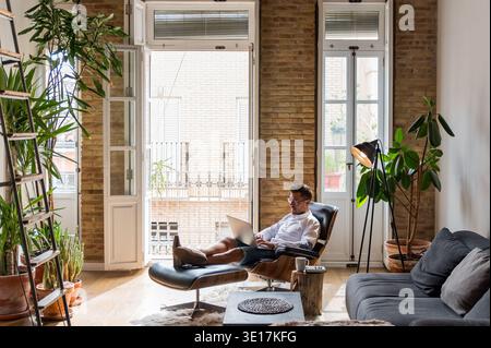 Handsome male entrepreneur sitting in armchair at home and working on project on laptop while enjoying coffee Stock Photo