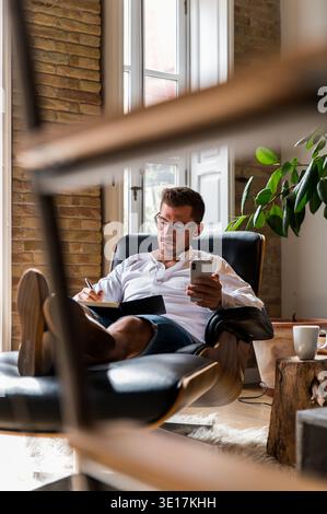 Focused male entrepreneur sitting in comfortable armchair and taking notes while planning working day at home office Stock Photo