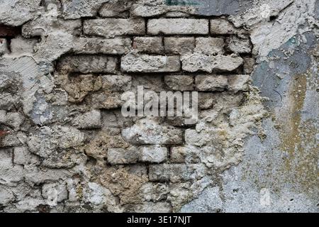 Old damaged brick wall with peeling grey plaster. Weathered urban background showing crumbling mortar and industrial grunge texture Stock Photo