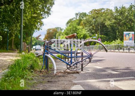 Old damaged blue bicycle abandoned and locked to a metal bike rack on a Paris street, missing front wheel, bent rear wheel with rusted spokes Stock Photo