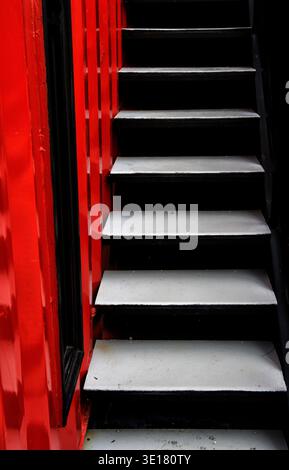 Modern black and white industrial staircase situated next to a vibrant red corrugated metal container wall. Stock Photo