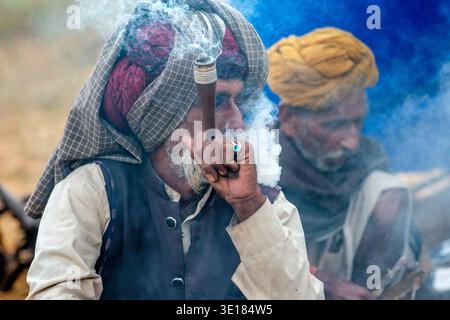 A man in a checked turban exhales smoke while using a traditional pipe at the Pushkar Camel Fair with another man seated in the hazy distance. Stock Photo