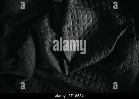 Close-up of black quilted bedsheet fabric in dramatic low light. Textured textile surface with deep shadows highlighting stitching pattern and soft fo Stock Photo