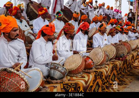 Large groups of men in white uniforms and orange turbans play traditional drums while seated on a patterned carpet during Pushkar Mela. Stock Photo