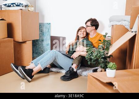 Couple sitting among moving boxes using laptop and celebrating relocation in new apartment interior Stock Photo