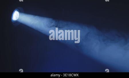 Blue spotlight beam shining through dense smoke on a dark stage. Theatrical lighting effect. Stock Photo