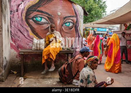 A man in saffron robes eats a banana on a bench in Pushkar in front of a mural of a woman’s face, with a crowded market alley in the distance. Stock Photo