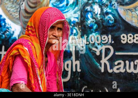 In Pushkar an elderly woman wears a vibrant pink and orange veil while sitting in front of a blue painted wall with white lettering and patterns. Stock Photo