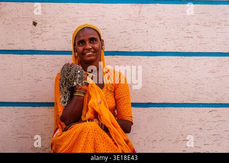 In Pushkar a smiling woman in a yellow dotted sari sits against a white and blue striped wall, holding a large bundle of silver-coloured necklaces. Stock Photo