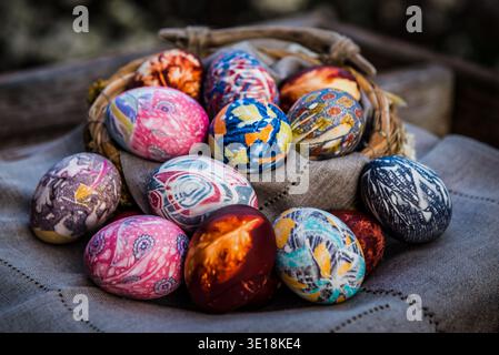 A collection of decorative silk-dyed Easter eggs nestled in a rustic woven basket on a linen cloth. Stock Photo