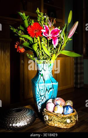 Festive Easter still life featuring silk-dyed eggs in a rustic basket beside a vibrant blue vase of lilies and gerberas. Stock Photo