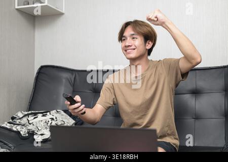 Young Thai Asian man wearing a brown t-shirt, sitting on a black leather sofa while using a laptop at home concept of modern lifestyle, remote work, s Stock Photo