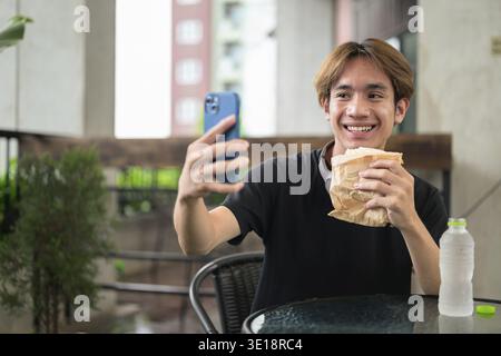 Happy young Asian Thai student man eating sandwich outdoors for casual lunch while sitting and using phone Stock Photo