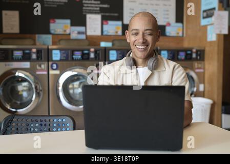 Bald Hispanic man in casual clothes sitting in laundry shop using laptop computer. Everyday urban lifestyle concept with washing machines in backgroun Stock Photo